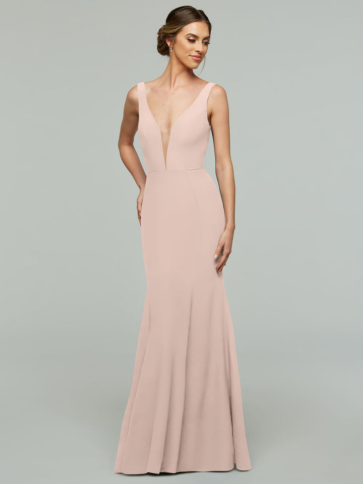 A woman in the Avery Austin 83010, a rose pink, floor-length mermaid dress with a deep plunging neckline, stands against a plain light gray background. Her hair is styled in an elegant updo.