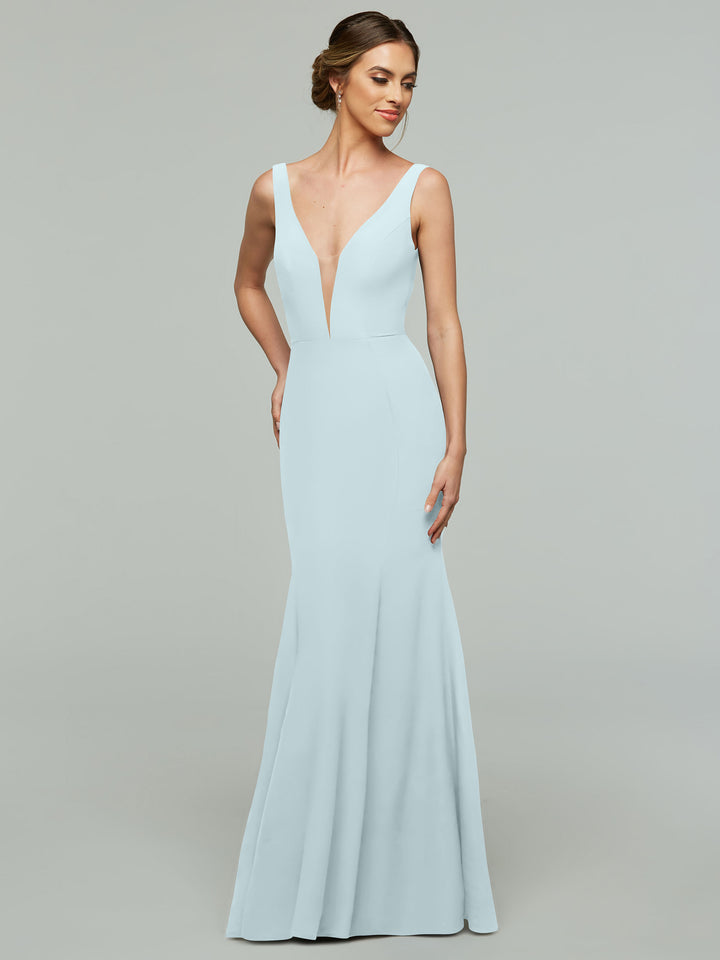 A woman in the Avery Austin 83010 sky blue sleeveless mermaid dress with a plunging neckline stands against a gray background, looking slightly to the side and smiling.