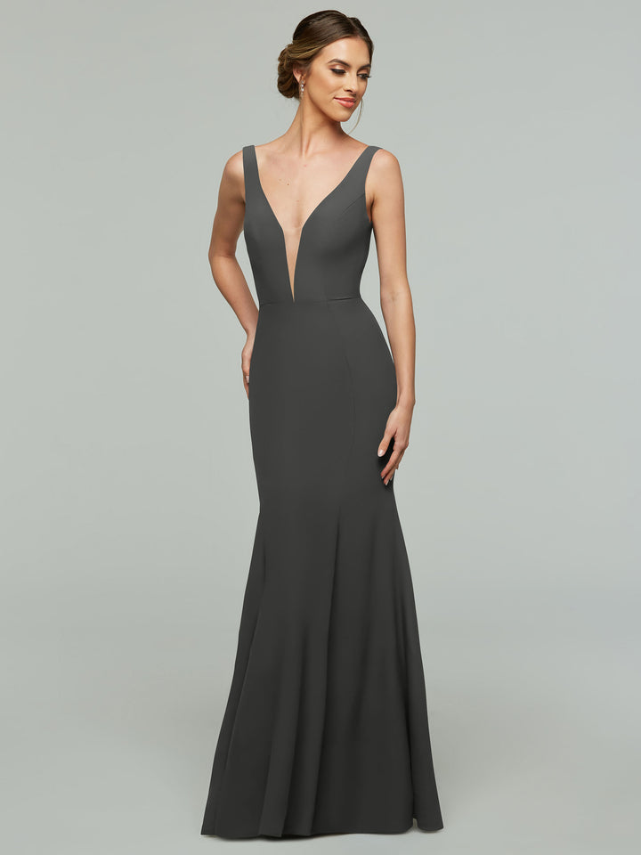 A woman models Avery Austin’s sleeveless, floor-length mermaid dress (style 83010) in Steel Gray, featuring a deep V neckline, as she stands against a light gray background and smiles gently to the side.