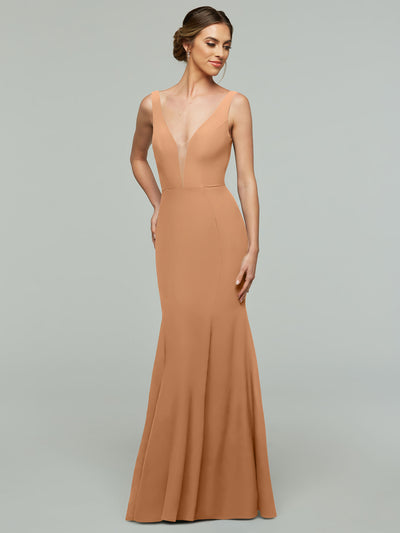 A woman in Avery Austin’s 83010, a sleeveless floor-length warm beige mermaid dress with a plunging neckline, stands against a plain light gray background. Her hair is styled in an elegant updo.