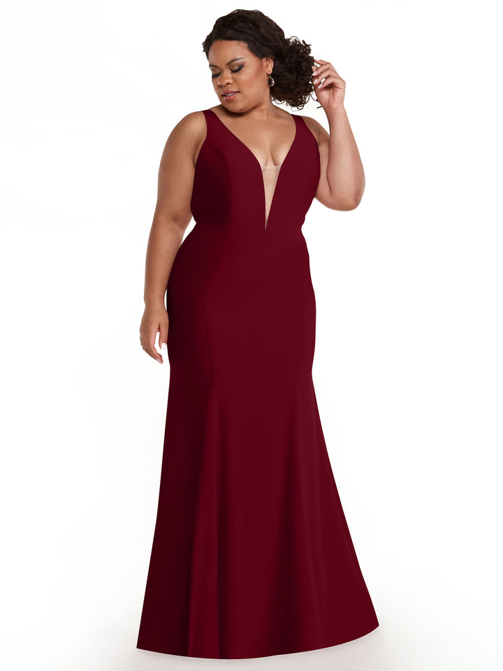 A woman wearing the Avery Austin 83010 burgundy sleeveless mermaid dress, featuring a deep plunging neckline and floor-length design, stands against a white background, looking down and adjusting her curly hair.