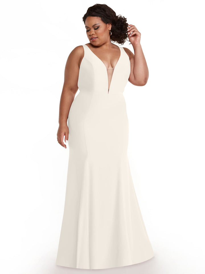 A woman in Avery Austin’s 83010, a floor-length white mermaid dress with a plunging neckline, stands against a plain white background, gazing to the side and touching her curly hair.