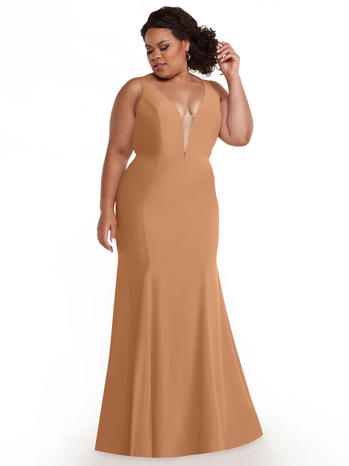 A woman models Avery Austin’s 83010, a sleeveless floor-length gown in warm beige with a plunging neckline, standing against a white background while looking down and touching her curly hair.