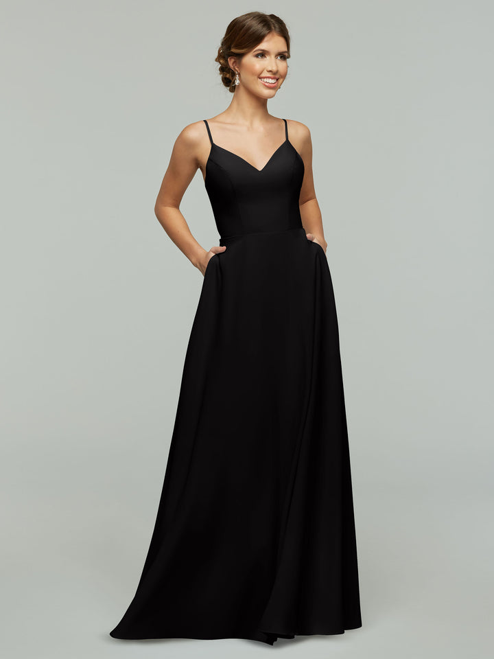 A woman stands smiling with her hands in the pockets of the Avery Austin 83012, a long black A-line dress with spaghetti straps and a V-neckline, against a plain light gray background.