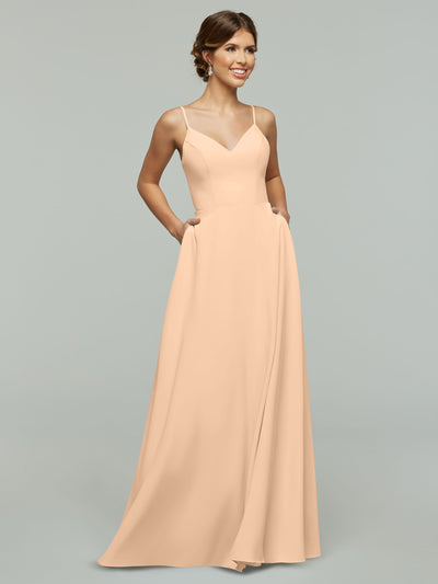 A woman smiles with her hands in the pockets of Avery Austin’s 83012—a light peach, floor-length A-line dress featuring a V-neck and thin spaghetti straps—posed against a plain gray background.