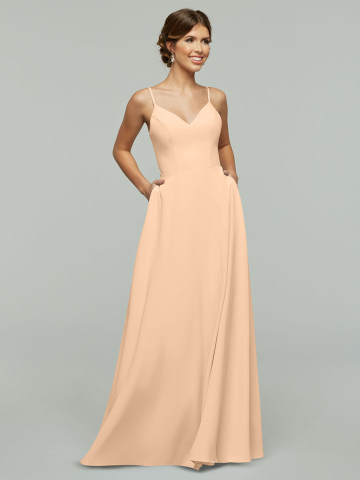 A woman smiles with her hands in the pockets of Avery Austin’s 83012—a light peach, floor-length A-line dress featuring a V-neck and thin spaghetti straps—posed against a plain gray background.