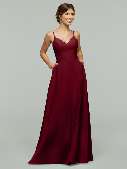 A woman smiles against a plain light gray background, modeling Avery Austin's 83012—a sleeveless, floor-length burgundy A-line dress with V-neck spaghetti straps and side pockets.