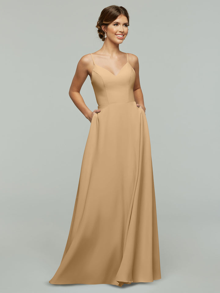 A woman in the Avery Austin 83012 A-Line dress, color Cappuccino, featuring a V-neck, spaghetti straps, and pockets, stands smiling against a plain light gray background.