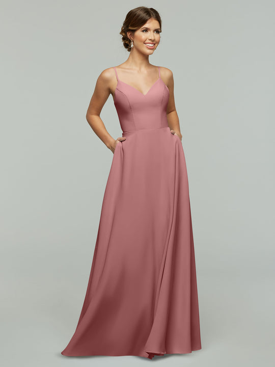 A woman wearing Avery Austin's 83012—an A-line, rose floor-length dress with spaghetti straps, V-neckline, fitted bodice, and pockets—stands smiling against a light gray background with her hair styled up.