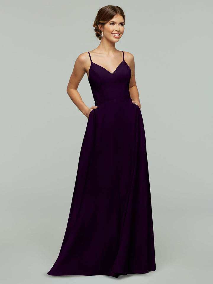 A woman models Avery Austin style 83012, a sleeveless floor-length deep purple A-line dress with spaghetti straps, V-neck, and pockets, smiling against a plain gray background.