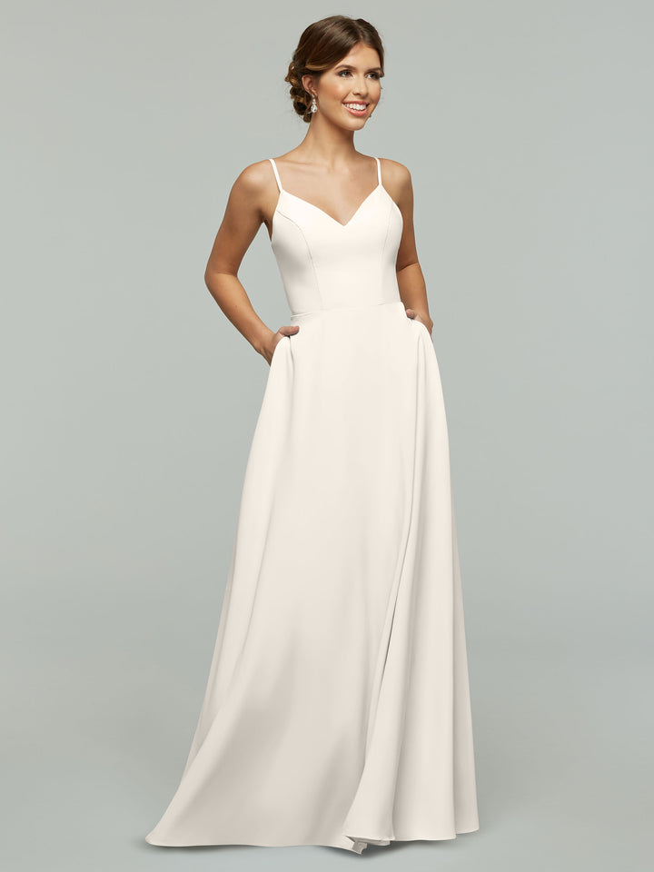 A woman in Avery Austin’s 83012 wears a sleeveless, floor-length white A-line dress with spaghetti straps and pockets, standing against a plain light gray background, smiling and looking slightly to the side.