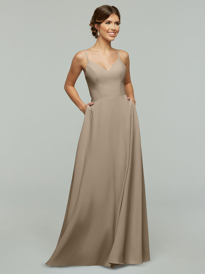 A woman models Avery Austin’s 83012—a long taupe A-line dress with a fitted bodice and spaghetti straps, featuring pockets—posing against a plain light gray background.