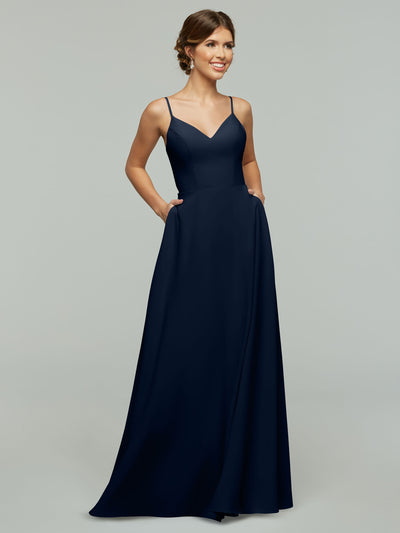 A woman stands smiling against a plain light gray background, wearing Avery Austin's 83012: a sleeveless, floor-length navy blue A-line dress with spaghetti straps, a fitted bodice, and pockets.