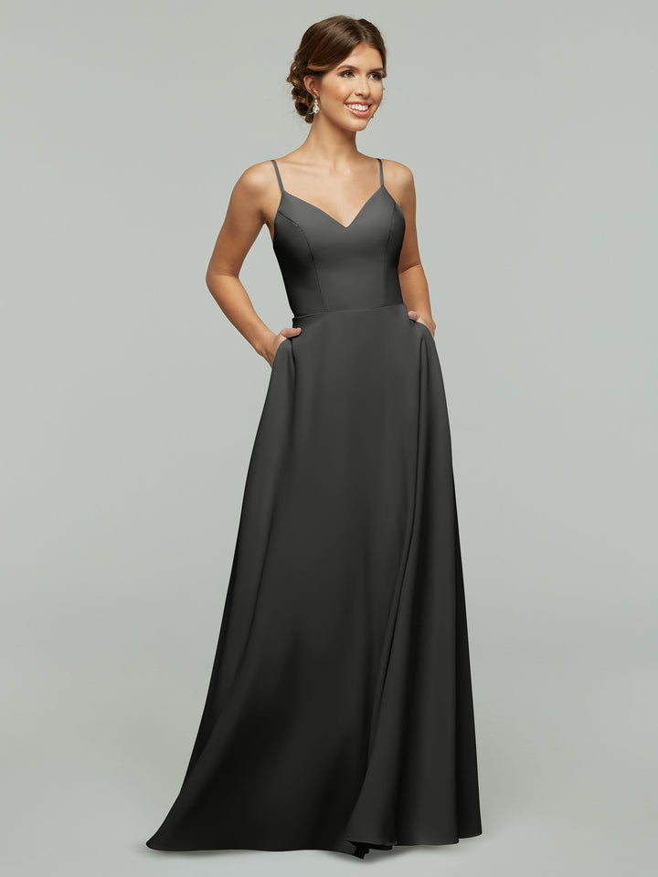 A woman smiles in the Avery Austin TH-83012, a sleeveless black A-Line gown with spaghetti straps, fitted bodice, pockets, and a flowing skirt. Her brown hair is styled up and she accessorizes with earrings.