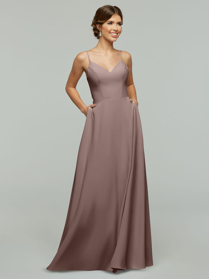 A woman stands smiling with her hands in the pockets of Avery Austin’s mauve, floor-length A-Line dress (Style 83012) featuring thin spaghetti straps against a light gray background.