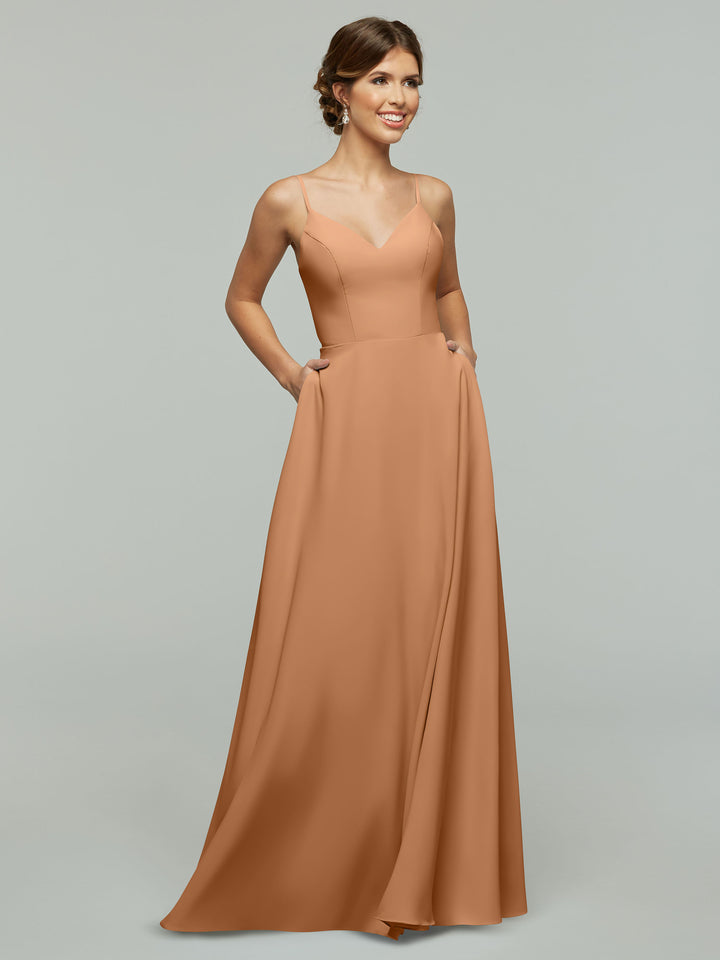 A woman smiles and looks to the side while wearing the Avery Austin TH-83012: a long, sleeveless light brown A-line dress with spaghetti straps, V-neck, and pockets, standing against a plain light gray background.