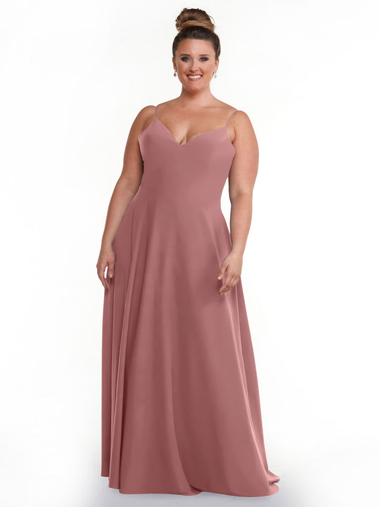 A smiling woman with her hair in a bun wears the Avery Austin TH-83012, a floor-length, sleeveless dusty rose V-Neck A-Line dress, standing against a plain white background.