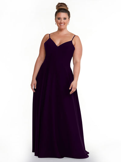 A smiling woman with brown hair in a bun models the Avery Austin 83012, a sleeveless, floor-length dark purple A-line dress with spaghetti straps and a V-neckline, against a plain white background.