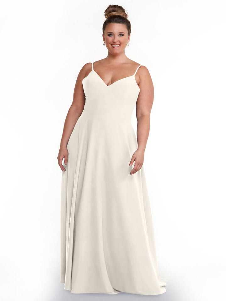 A woman stands and smiles against a plain white background, wearing the Avery Austin 83012: a sleeveless, floor-length white A-line dress with spaghetti straps and a V-neckline.