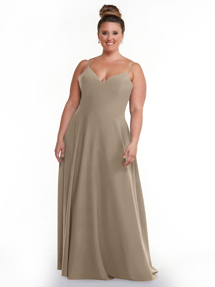 A smiling woman with her hair in a bun models Avery Austin’s floor-length taupe A-line dress (style 83012) with spaghetti straps and a V-neckline, standing against a plain white background.