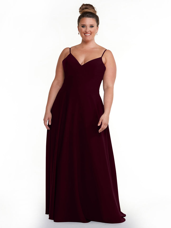 A woman with a high bun smiles in front of a white background while wearing Avery Austin’s 83012, a sleeveless, floor-length deep burgundy A-Line dress with spaghetti straps.