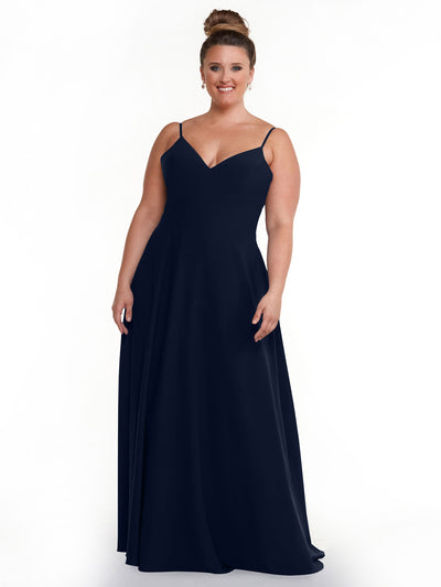 A woman with her hair in a bun wears Avery Austin’s 83012, a navy blue floor-length A-line dress with spaghetti straps and a V-neckline, smiling while standing against a plain white background.