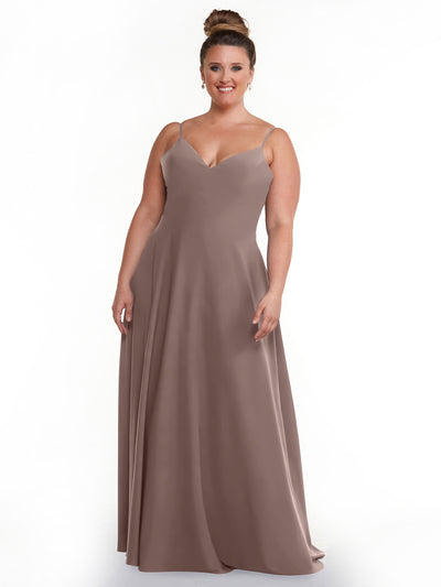 A woman stands smiling against a white background wearing Avery Austin style 83012, a mauve floor-length A-line dress with spaghetti straps. Her hair is in a high bun and she’s accessorized with earrings.