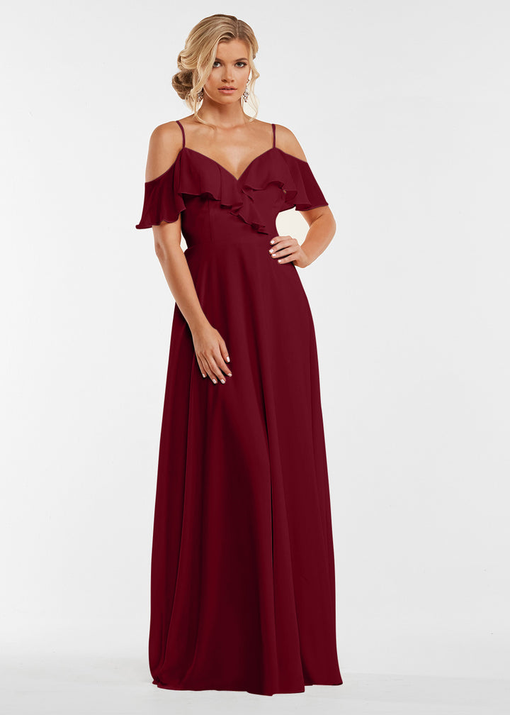 A woman models Avery Austin’s 83013, a floor-length burgundy gown with off-the-shoulder sleeves and ruffled bodice, standing against a white background with her hair in soft waves.