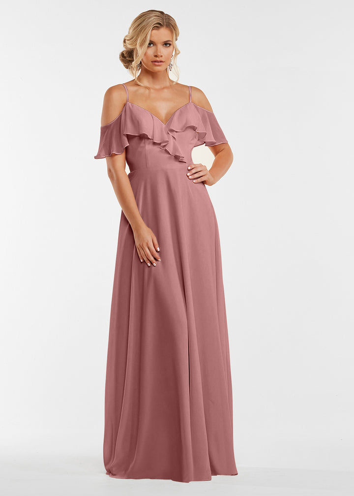 A woman models the Avery Austin TH-83013 gown in dusty rose, featuring off-the-shoulder sleeves and ruffled bodice detail. She stands against a plain white background with her hair styled in loose curls.