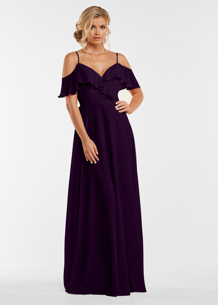 A woman models Avery Austin’s 83013—a floor-length, deep purple gown with a ruffled neckline and off-the-shoulder sleeves—standing against a plain white background, her hair in loose waves as she gazes forward in a relaxed pose.