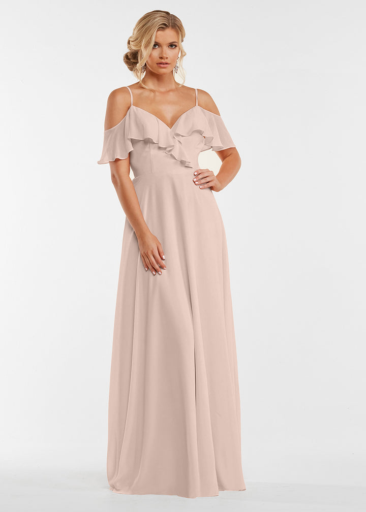 A woman wears Avery Austin’s 83013, a long rose pink dress with off-the-shoulder sleeves and ruffled bodice, standing against a plain white background with her hair in loose waves.