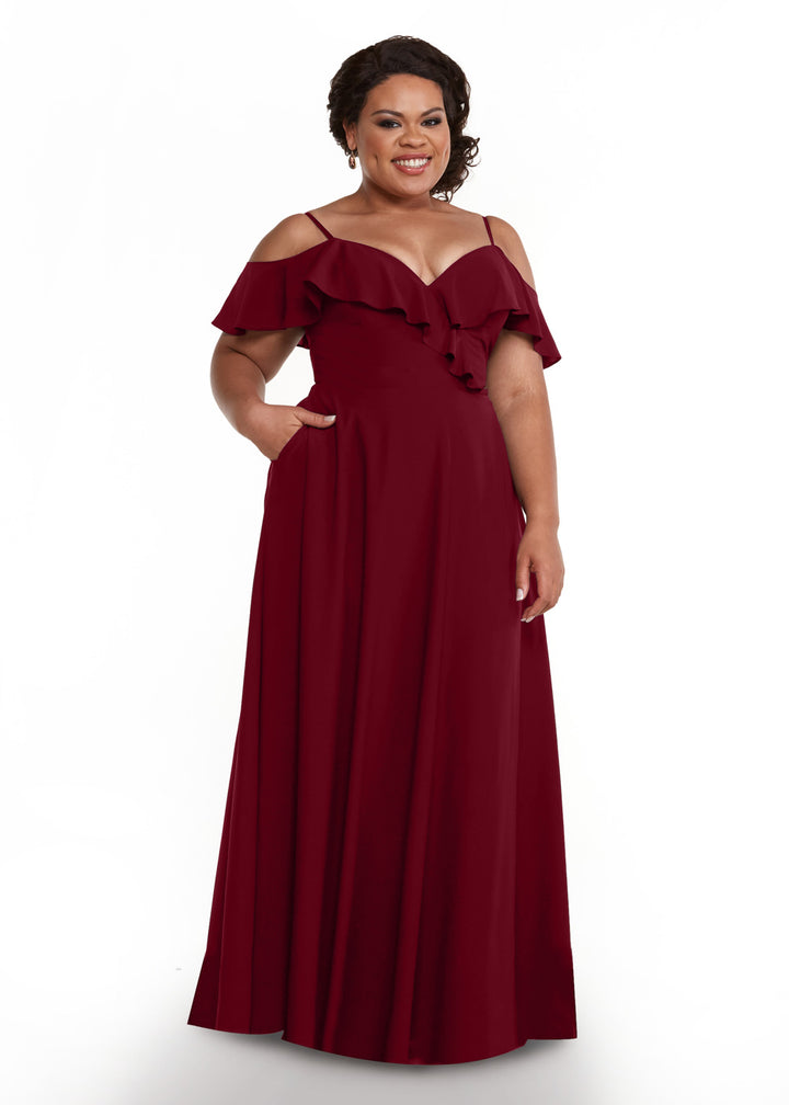 A woman smiles with one hand in her pocket, modeling the Avery Austin 83013—a floor-length burgundy dress featuring off-the-shoulder sleeves and ruffle details—against a white background.