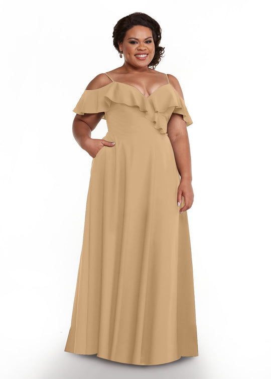 A woman stands smiling against a white background, wearing Avery Austin’s 83013: a light brown, floor-length dress with ruffled neckline, off-the-shoulder sleeves, and delicate spaghetti straps.