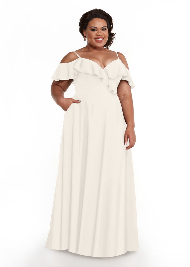 A woman with dark, curly hair models Avery Austin’s 83013: a floor-length white dress with a ruffled neckline and spaghetti straps, standing with one hand in her pocket against a plain white background.