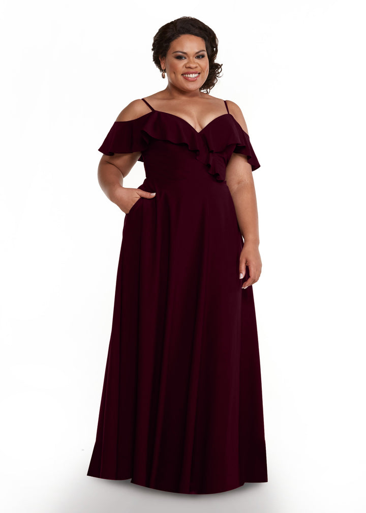 A woman wearing the Avery Austin 83013, a floor-length burgundy dress with off-the-shoulder sleeves, spaghetti straps, and a ruffled neckline, stands smiling with one hand in her pocket against a plain white background.