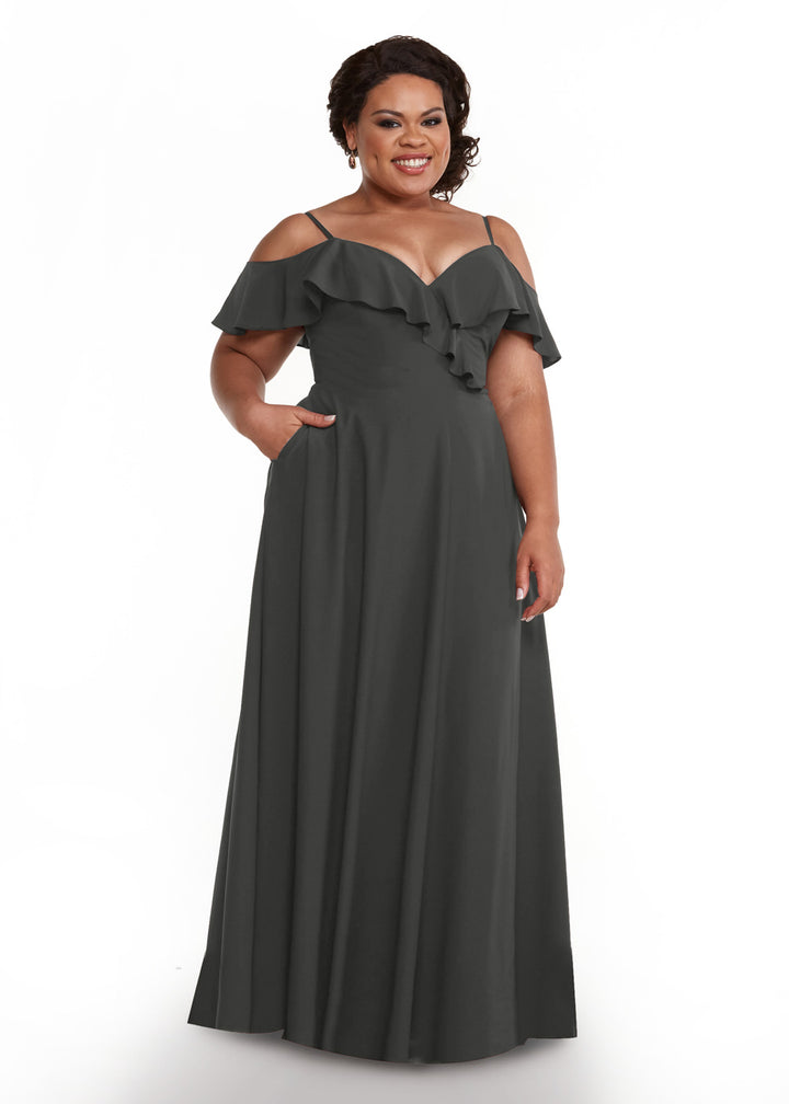 A woman models Avery Austin’s 83013: a dark, floor-length, off-the-shoulder gown with a ruffled neckline. She stands smiling with one hand in her pocket against a white background.