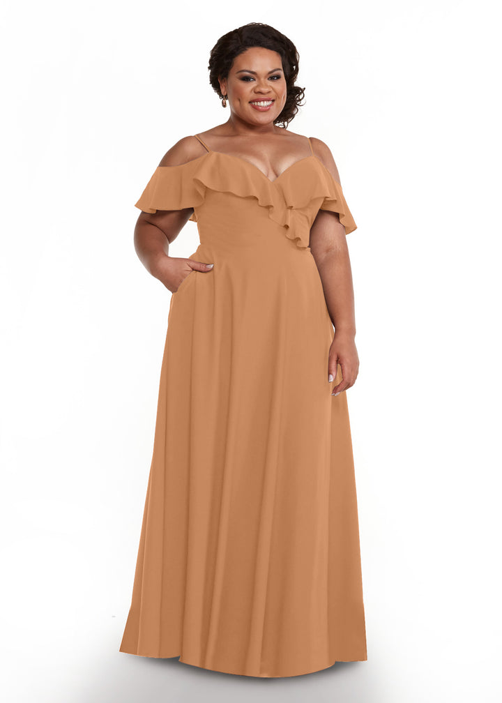 A woman with short curly hair smiles while wearing the Avery Austin 83013 long, off-the-shoulder light brown ruffled dress with a ruffled neckline and short sleeves, standing against a plain white background.