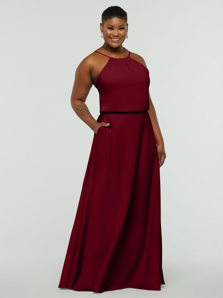 A person with short hair smiles while wearing Avery Austin’s 83015: a dark red, sleeveless, floor-length A-Line dress with a fitted bodice, flowing skirt, and convenient pockets. They stand with one hand in a pocket against a light background.