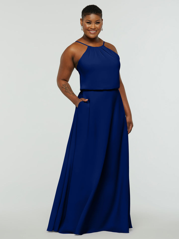 A person stands smiling in Avery Austin’s 83015: a sleeveless, floor-length royal blue A-Line dress with pockets, a high neckline, and subtle pleating. The background is plain and light-colored.