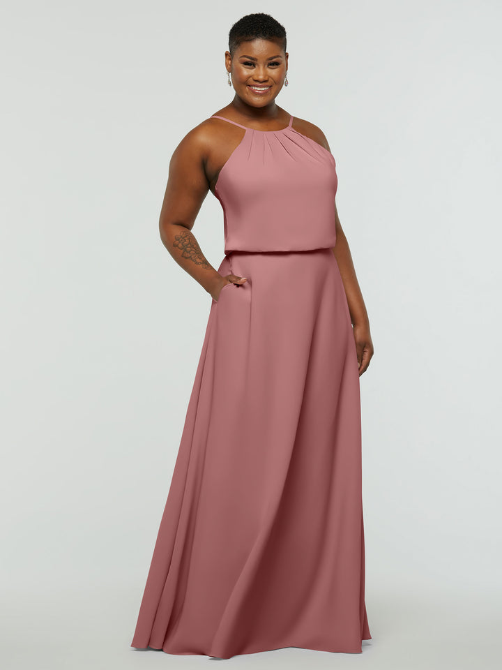 A person stands smiling with one hand in their pocket, wearing the Avery Austin TH-83015 sleeveless, floor-length dusty rose A-line dress with pleated halter neck detailing, against a plain light background.