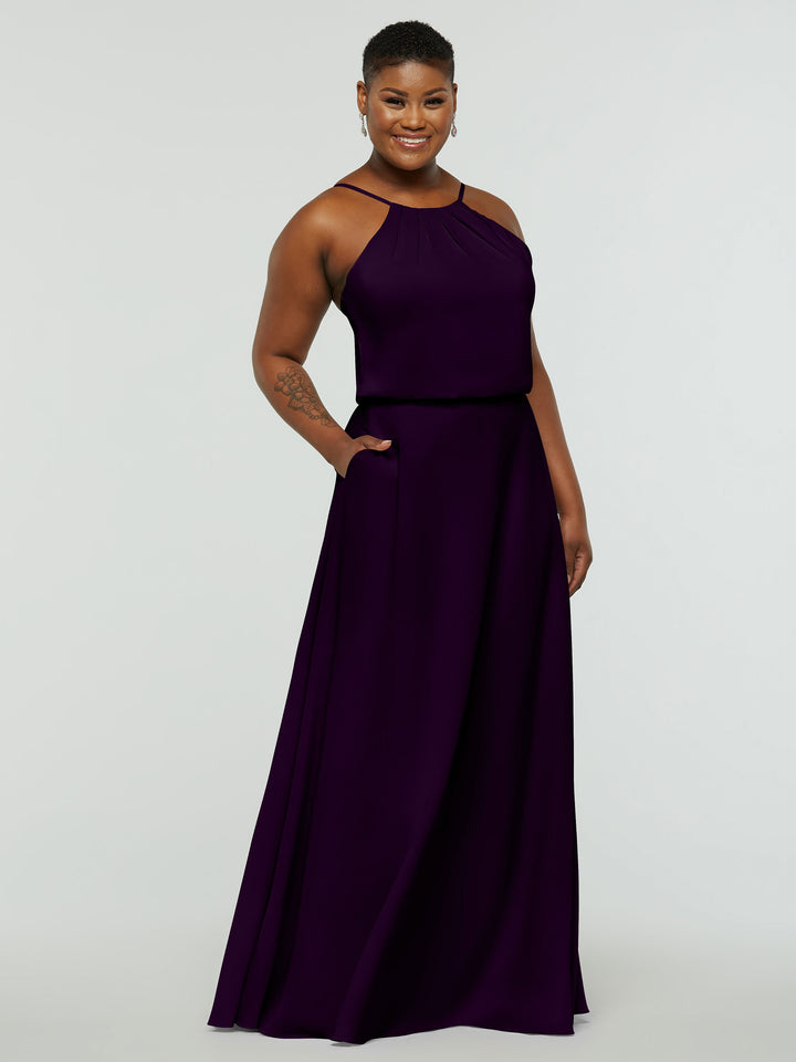 A person stands smiling in a sleeveless, floor-length Avery Austin 83015 dark purple A-line dress with pockets, high neckline, and subtle pleating. The background is plain and light-colored.