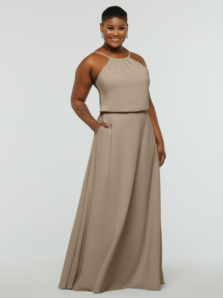 A woman with short hair smiles in Avery Austin’s 83015— a sleeveless, taupe A-line dress with pockets, high pleated neckline, and fitted waistband—against a plain white background.