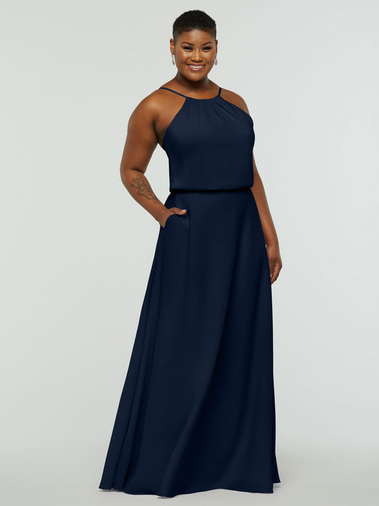A person stands smiling with one hand in the pocket of the Avery Austin 83015 sleeveless, floor-length navy blue A-line dress with a halter neckline against a plain light background.