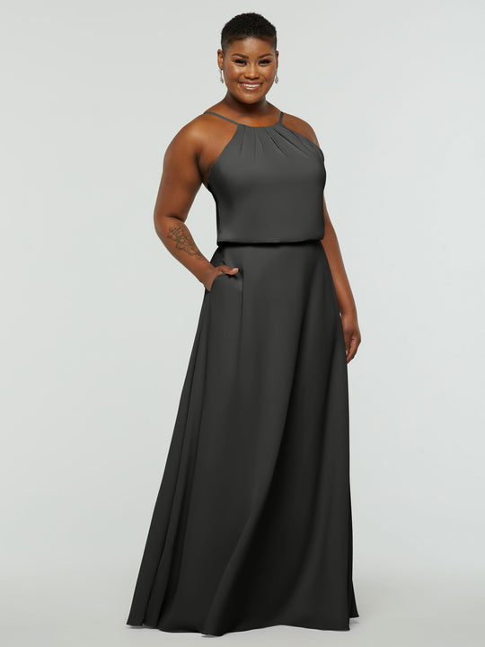 A person smiles in the Avery Austin 83015, an elegant sleeveless floor-length dark gray A-line dress with a halter neck, pleated neckline, and pockets, standing against a plain light background.