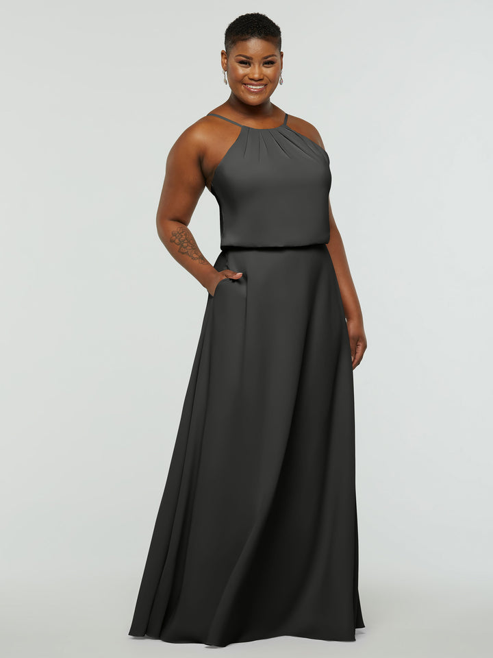 A person smiles in the Avery Austin 83015, an elegant sleeveless floor-length dark gray A-line dress with a halter neck, pleated neckline, and pockets, standing against a plain light background.