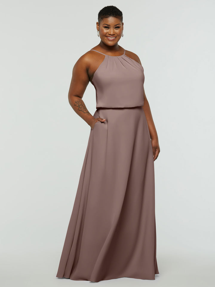A woman smiles in the Avery Austin 83015, a sleeveless, floor-length mauve A-Line dress with a halter neck and pleated details. She stands with one hand in her pocket against a plain white background.