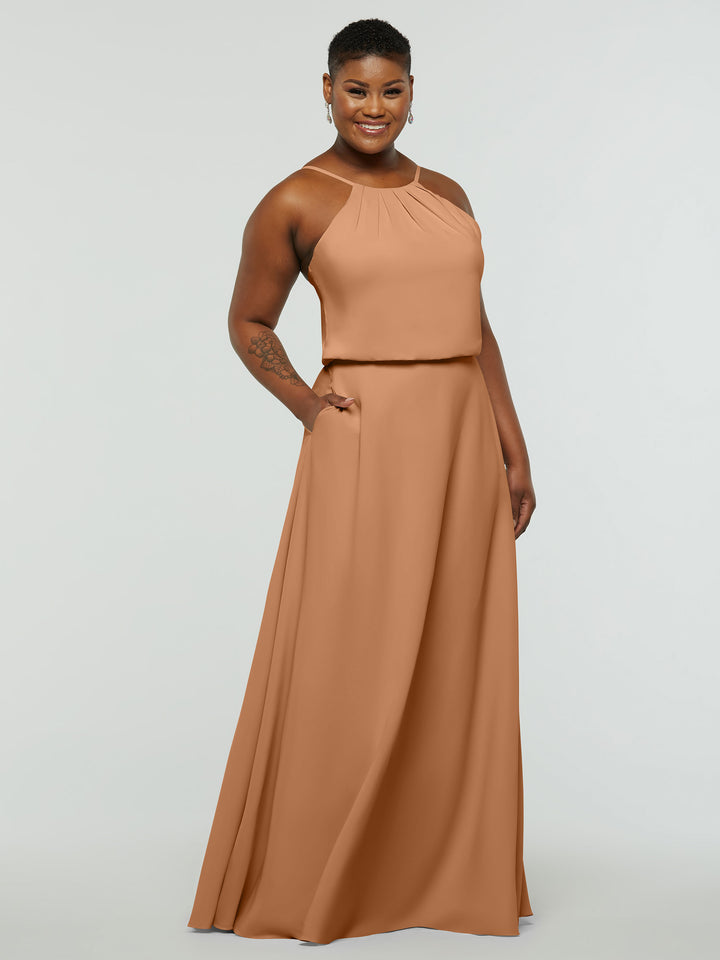 A person wearing Avery Austin’s 83015, a sleeveless floor-length A-Line dress with a halter neck in light brown, stands smiling with one hand in their pocket against a plain light background.