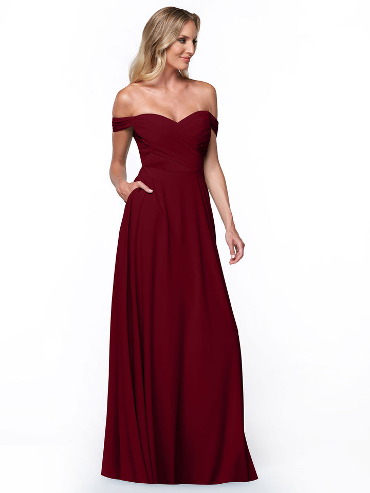 A woman with blonde hair stands against a white background in Avery Austin’s 83018: a long, off-the-shoulder dress featuring pockets and an elegant A-line skirt, with one hand casually placed in her pocket as she gazes to the side.