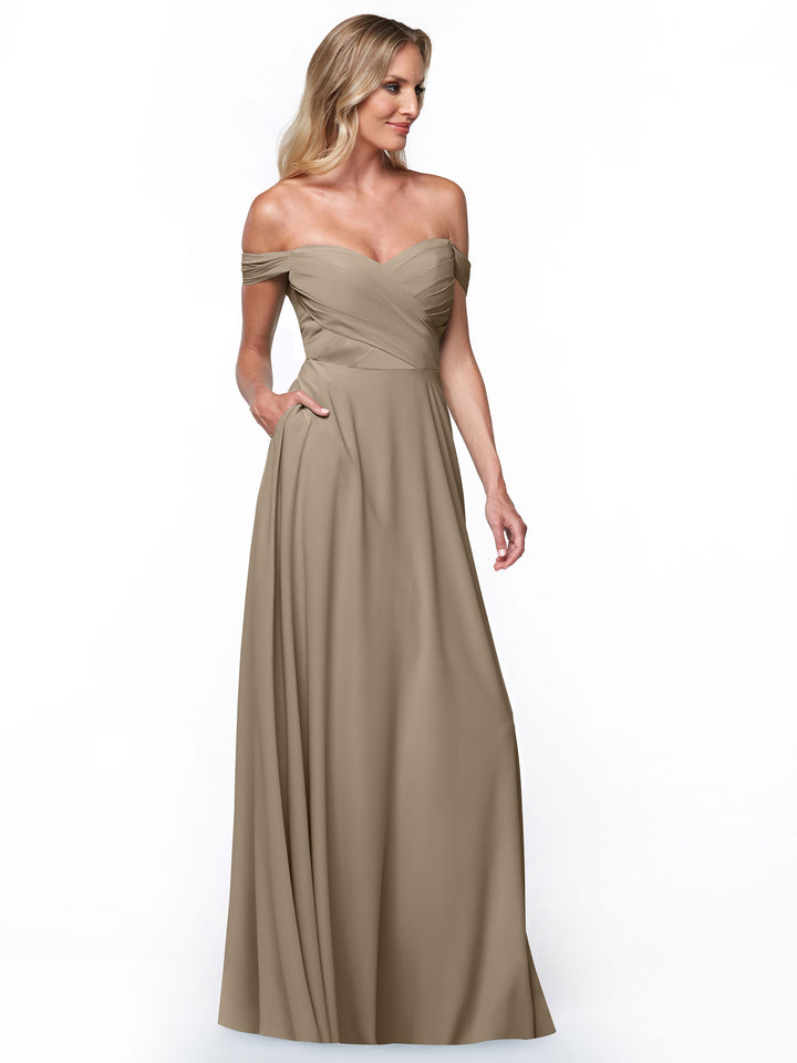 A woman models the Avery Austin 83018 gown, showcasing its off-the-shoulder fitted bodice, pleated details, and elegant A-line skirt. Posed against a white background, she highlights the gown’s taupe color and graceful silhouette.