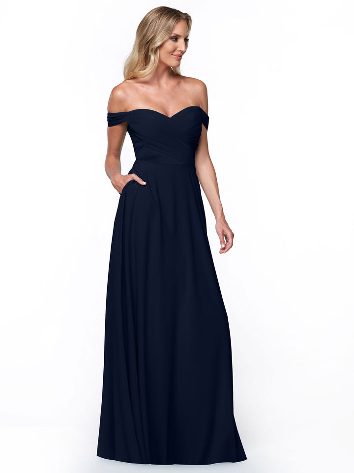 A woman models Avery Austin’s 83018, a navy blue off-the-shoulder gown with pockets, a fitted pleated bodice, and a flowing A-line floor-length skirt, set against a crisp white background.